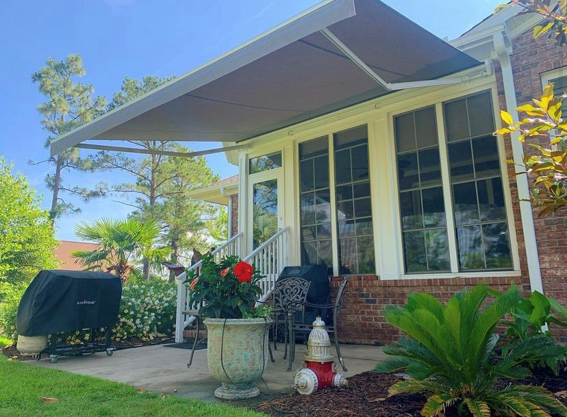 Gray retractable awning covering a patio on a classic brick home surrounded by tropical landscaping including palm trees and hibiscus flowers